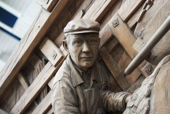 A relief in lime: a man in a flat cap and work jacket leans out from stacked timber and sawmill framing cut in the same block.