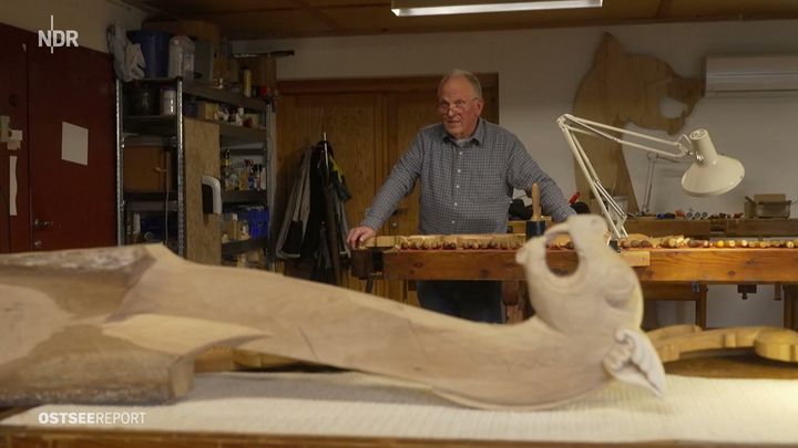 Rolf stands at the workbench in his workshop, hands resting on it. In the foreground lies a long, half-carved stem in pale wood ending in a curled head, and behind him a large template of a dragon head hangs on the wall.