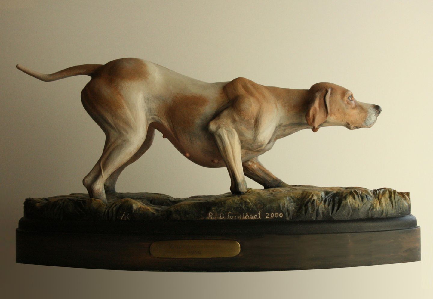 A pointer carved in birch and painted brown and white, standing on point on a base of grass.
